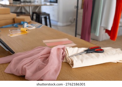 Sewing studio worktable showing soft pink chiffon draping beside cream fabric bolt and scissors. Textile, swatch, cloth, wooden, workshop, garment, rack