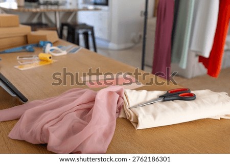 Sewing studio worktable showing soft pink chiffon draping beside cream fabric bolt and scissors. Textile, swatch, cloth, wooden, workshop, garment, rack