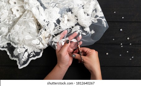 Sewing wedding dress. Hands of woman seamstress tailor sewer dressmaker designer holding scissors and cutting white lace and sew beads on a dark wooden background. Top view. Horizontal orientation. - Powered by Shutterstock