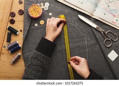 Sewing workshop or fashion designer at work. Top view on female hands marking cloth square with soap piece and measuring tape. Messy table with dressmaker accessories and patterns, copy space