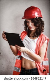 Sex Equality And Feminism. Sexy Girl In Workwear Safety Helmet And Orange Vest Using Tablet Touchpad Ipad On Gray. Young Attractive Woman As Construction Worker. Technology.