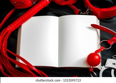 Sexual Kink And BDSM Concept Theme With An Open Book Or Notebook With Blank White Pages Sounded By Handcuff, Whip, Flogger, Collar And Leash, Ball Gag And Other Bondage Devices With Copy Space