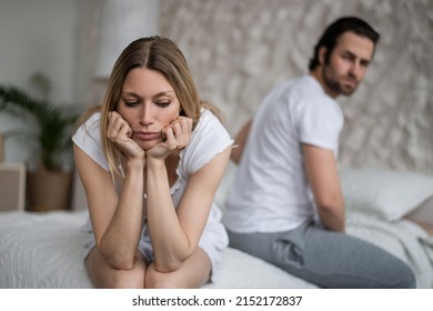 Sexual Problems, Relationship Crisis Concept. Stressed Young Woman Sitting On Bed, Overwhelmed With Family Difficulties, Her Offended Husband On Background, Blank Space