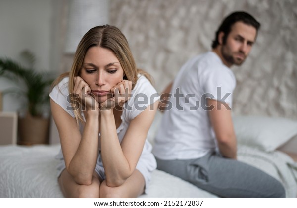 Sexual problems, relationship\
crisis concept. Stressed young woman sitting on bed, overwhelmed\
with family difficulties, her offended husband on background, blank\
space