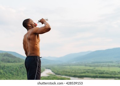 Sexy african american muscular man drinking water after sports training. Breathtaking green mountain landscape on background