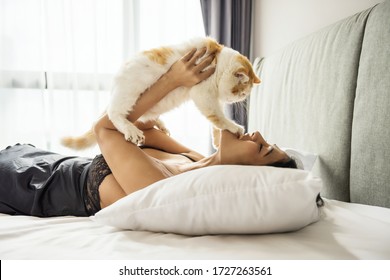 Sexy Asian Woman In Black Pajama Dress Play With Yellow Exotic Shorthair Caton White Bed. Stay Home During Covid-19 Pandemic Disease. WFH Or Work From Home, Quarantine, Social Distancing.