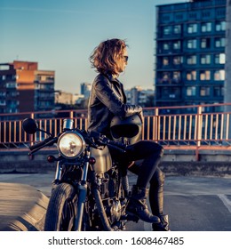 Sexy biker beautiful female in black trendy leather jacket sit on vintage custom made caferacer motorcycle. Urban roof parking, sunset in big city. Traveling and active hipster lifestyle. Girls power.