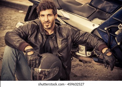 Sexy Biker Man Wearing Jeans And Leather Jacket Sitting Relaxed By His Motorcycle.