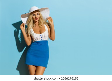 Sexy Blond Woman In Blue White Mini Dress And Sun Hat Is Looking At Camera. Three Quarter Length Studio Shot On Turquoise Background.