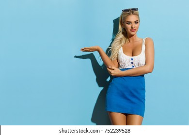 Sexy Blond Woman In Blue White Mini Dress Is Gestures And Presenting. Three Quarter Length Studio Shot On Turquoise Background.