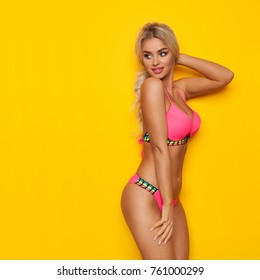 Sexy blond woman in pink bikini is looking over the shoulder at copy space. Three quarter length studio shot on yellow background