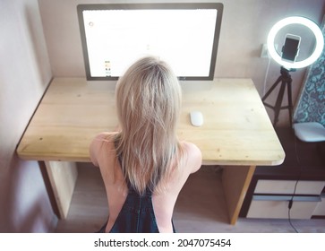 Sexy Blonde Woman In Denim Jumpsuit Sitting At A Table Near A Laptop And A Smartphone In Online Video Chat. Web Model Concept.