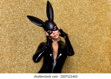 Sexy Blonde Woman Posing In Halloween Vinyl Black Costume And Black Bunny Mask On On A Glitter Gold Background. Halloween Costume.