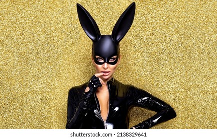 Sexy blonde woman posing in latex costume and black bunny mask on gold glitter background. Easter bunny concept. Latex lingerie. Naughty girl. Halloween costume.