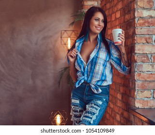 A Sexy Brunette In A Flannel Shirt And Jeans Holds A Cup Of Coffee Posing Against A Brick Wall. 