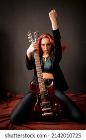 Sexy And Brutal Rocker Punk Or Metal Red-hair Girl With An Electric Guitar In A Black Jacket And Jeans Is Sitting On The Floor On Her Knees In A Defiant Pose And Hand Up In The Air