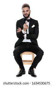 Sexy Businessman Wearing Black Tuxedo Sitting On A Wooden Chair And Making A Fuck You Sign Being Grumpy On White Studio Background