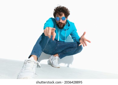 Sexy Casual Man Pointing At The Camera, Squatting And Wearing Sunglasses In A Fashion Pose