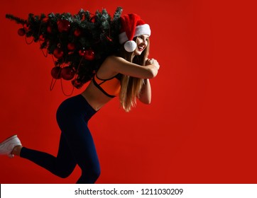 Sexy Christmas Fitness Sport Woman Wearing Santa Hat Holding  Xmas Tree On Her Shoulders. Running Forward On A Red Background