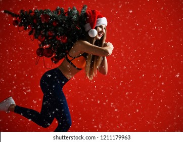 sexy Christmas fitness sport woman wearing santa hat holding  xmas tree on her shoulders. running forward on a snowing red background
