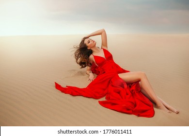 Sexy Elegant Stylish Woman Model Lies Posing On Summer Desert. Red Luxury Silk Trendy Long Dress. Beautiful Bare Legs. Orange Sand Dune Beach Sunset Beauty Evening Sky. Brunette Loose Hair Fly In Wind