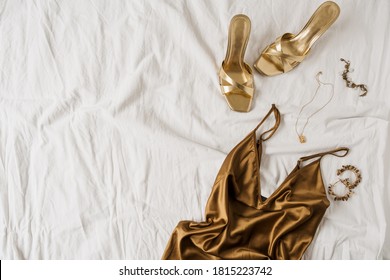 Sexy fashion concept with women's clothes and accessories. Golden shoes, brown silk dress, golden necklace, bracelet, earrings on white linen. Flat lay, top view minimal feminine vogue background.