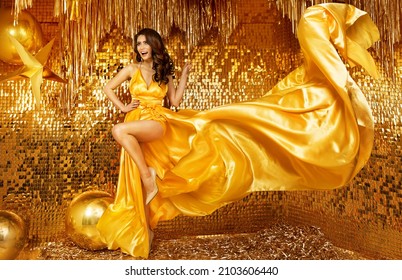 Sexy Fashion Model in Golden Silk Dress Dancing with flying Fabric in Gold Sparkling Interior. Beauty Glamour Woman in luxury long Yellow Gown