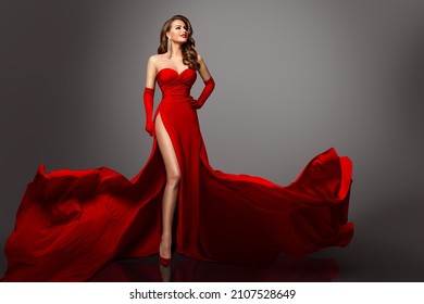 Sexy Fashion Model in Red Silk Dress and Gloves. Glamour Woman in Long Luxury Slit Gown flying on Wind with Wavy Hair Style showing Leg over Gray Background