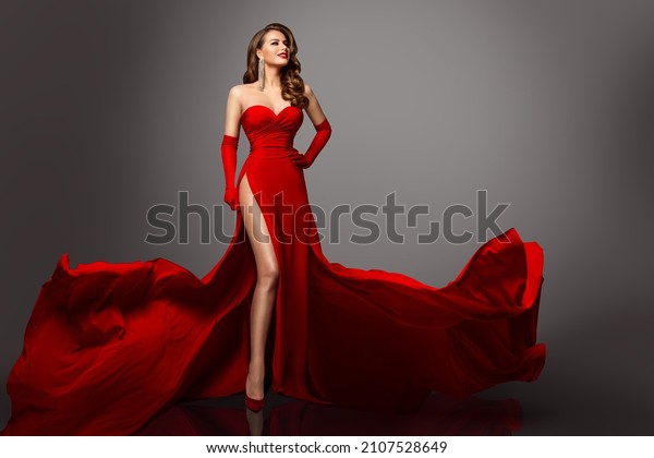 Sexy Fashion Model in Red Silk Dress\
and Gloves. Glamour Woman in Long Luxury Slit Gown flying on Wind\
with Wavy Hair Style showing Leg over Gray\
Background