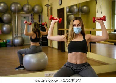 Sexy Female Athlete Fitness Coach Wearing White Medical Protective Mask Do Exercises With Red Dumbbell Invite To Do Online Fitness Class Sitting On Yoga Ball In Empty Gym. Athletes Against COVID-19