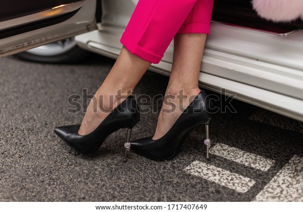 Sexy female elegant legs in luxurious stylish\
pink trousers in fashionable black leather high-heeled shoes in\
auto on parking. Trendy girl model in business clothes is resting\
in car. Close-up.