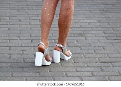 Sexy female legs on high heels on a street, slim woman in silver-white shoes and fishnet stockings with rhinestones. Concept of ladies fashion and footwear, elegance, summer weather