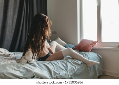 Sexy girl in black lingerie in the bed. Young brunette with long curly hair is sitting at the window in a man's shirt.