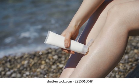 Sexy Girl On The Beach With Sunblock Or Suntan Cream. Sits On The Sand By The Sea On The Beach. Smear Cream On Arm, Shoulder And Legs.