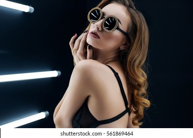 Sexy Girl Posing In Studio Against Black Background With Lamps, Wearing Sunglasses.Joyful Fashion Woman With Bright Make Up,red Lips In Sunglasses,black Panties And A Black Bra