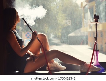 Sexy girl in t-shirt, short jeans and sneakers sitting at the window and smoking hookah. 
