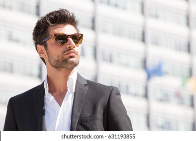 Sexy lindo homem elegante. Óculos de sol. Estilo da cidade. Um homem bonito e encantador com óculos de sol ao ar livre. Cabelo restolho e preto. Luz intensa. Camisa branca e jaqueta elegante.