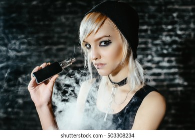 Sexy goth girl smokes electronic cigarette on dark background. The model vaper vaping a vaporizer in the studio.