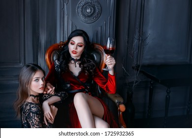 Sexy gothic woman vampire evil sitting on armchair holding glass of wine blood. bloody makeup sharp teeth fangs red lips. vintage medieval dress. frightened scared girl victim obeys at feet mistress