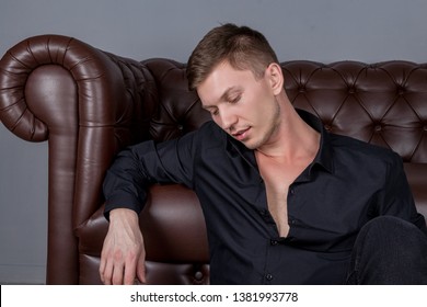 Sexy handsome man wearing black shirt sitting on floor near the leather sofa. Comfort and relaxation