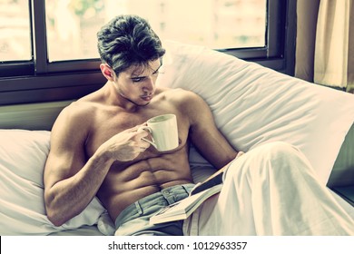 Sexy handsome young man laying shirtless on his bed next to window, holding a coffee or tea cup while reading a book