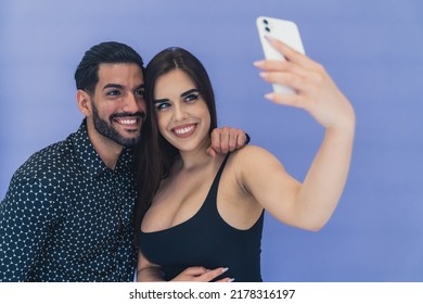 Sexy Heterosexual Couple Taking Selfie Over Violet Background In Studio. Two Smiling People Taking A Photo Of Themselves With A White Phone. High Quality Photo