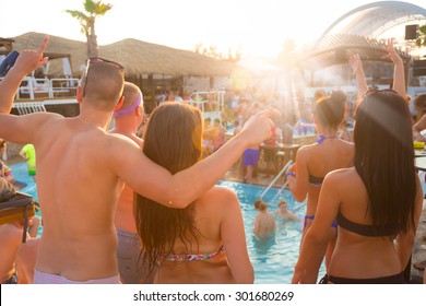Sexy hot friends dancing on a beach party event in sunset. Crowd dancing and partying at poolside in background. Summer electronic music festival. Hot summer party vibe.