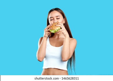 Sexy Hungry Athletic Woman With Closed Eyes Enjoying Eating Hamburger On Blue Background. Close Up Photo. Pleasure, Lifestyle, Food Studio Shot