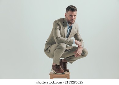 Sexy Man With Beard In Suit Looking Away While Crouching On Wooden Chair And Thinking About Work On Grey Background In Studio