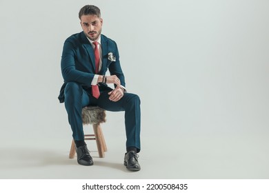 Sexy Man In Elegant Suit Holding Elbow On Knee And Sitting On Fur Chair In Front Of Grey Background In Studio