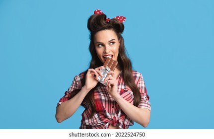 Sexy Millennial Pinup Woman In Retro Outfit Eating Chocolate Bar On Blue Studio Background, Panorama. Gorgeous Lady In Old Fashioned Wear Biting Delicious Sweet Dessert