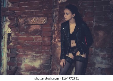 Sexy Model Stands Old Brick Wall And Poses In The Image Of A Corrupt Girl. Prostitute On The Street Smoking A Cigarette With His Knee Bent. Girl Addict In A Leather Jacket