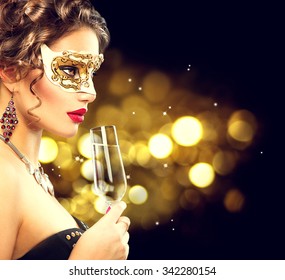 Sexy Model Woman With Glass Of Champagne Wearing Venetian Masquerade Mask At Party, Drinking Champagne Over Holiday Glowing Background. Christmas And New Year Celebration