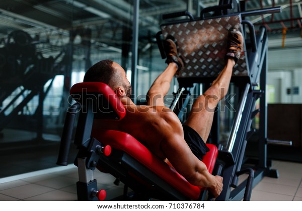 Sexy muscular men\
using a leg press machine and placing his legs on the platform on a\
dark colorful background.
